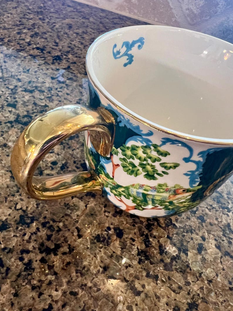 May include: A white ceramic teacup with a gold handle and rim. The cup features a detailed landscape scene with green trees and a colorful chameleon. Blue decorative accents are on the rim.
