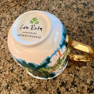 May include: White ceramic mug with a gold handle and a landscape design in green and blue. The mug has the text "Lou Rota exclusively for ANTHROPOLOGIE" printed on the bottom.