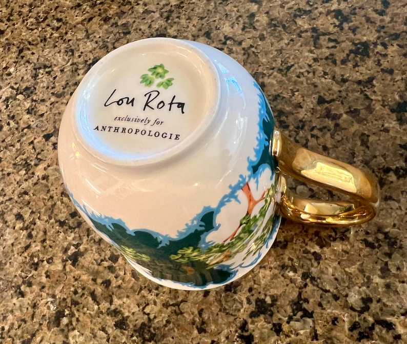 May include: White ceramic mug with a gold handle and a landscape design in green and blue. The mug has the text "Lou Rota exclusively for ANTHROPOLOGIE" printed on the bottom.