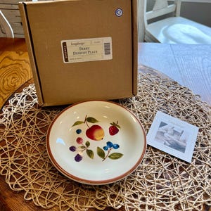 May include: A white ceramic dessert plate with a berry design, including a red apple, strawberries, and blueberries. The plate is next to its original cardboard box, labeled "Berry Dessert Plate."