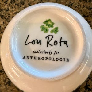 May include: White ceramic teacup base with the text "Lou Rota exclusively for ANTHROPOLOGIE" in black. The teacup base has a gold handle and a green floral design.