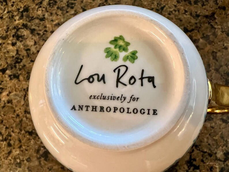 May include: White ceramic teacup base with the text "Lou Rota exclusively for ANTHROPOLOGIE" in black. The teacup base has a gold handle and a green floral design.
