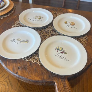 May include: A set of four white ceramic plates with thin black rims, each featuring a hand-drawn illustration of a specific cheese and its name written in black script: Brie, Camembert, Gouda, and Stilton, arranged on a round wooden board.