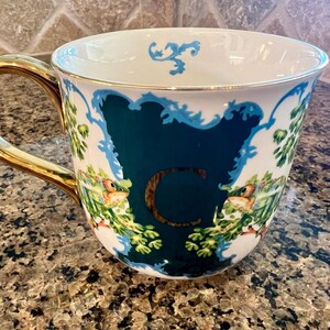 May include: White ceramic mug with a gold handle and rim. The mug features a teal-colored design with the letter "C" in gold, surrounded by floral and bird illustrations. The mug is on a speckled brown surface.