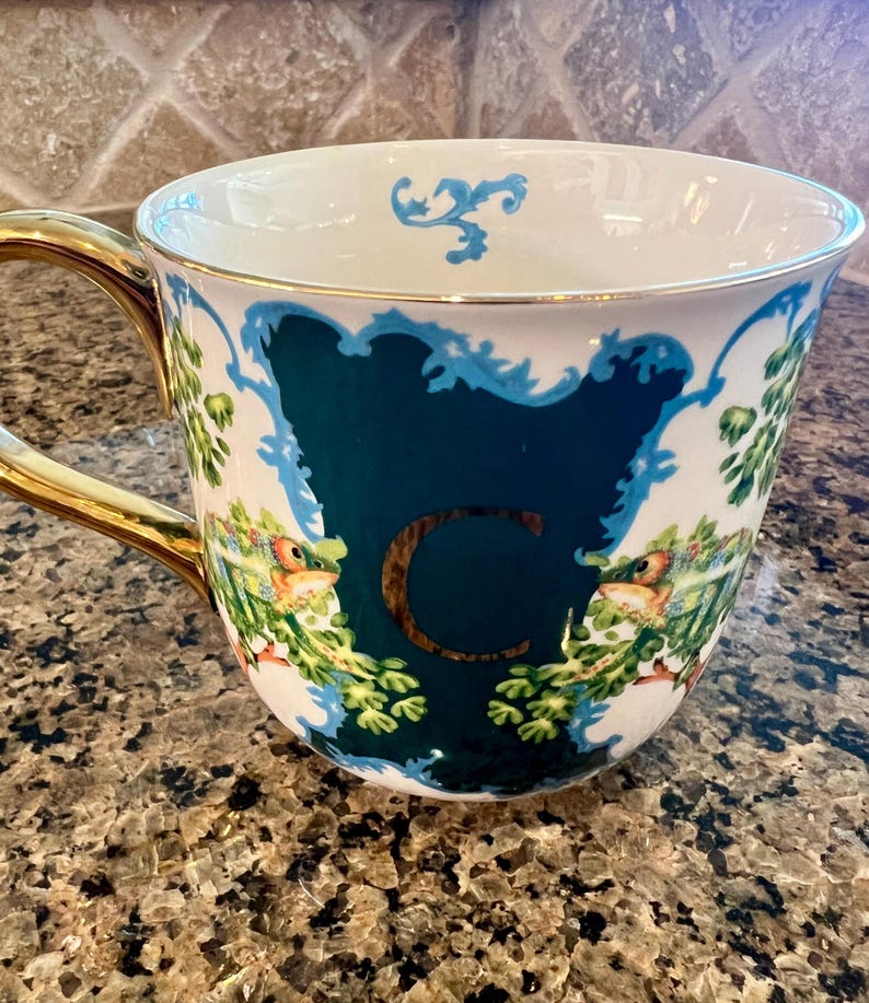 May include: White ceramic mug with a gold handle and rim. The mug features a teal-colored design with the letter "C" in gold, surrounded by floral and bird illustrations. The mug is on a speckled brown surface.