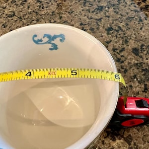 May include: A white ceramic bowl with blue floral accents, approximately 5 inches in diameter, is measured with a yellow tape measure. A red and black tape measure is also visible.