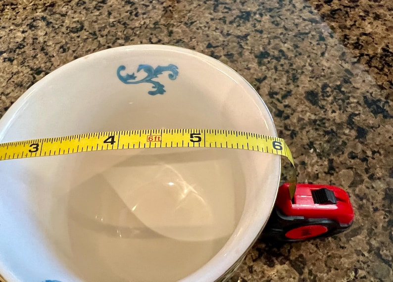 May include: A white ceramic bowl with blue floral accents, approximately 5 inches in diameter, is measured with a yellow tape measure. A red and black tape measure is also visible.