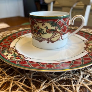 May include: A porcelain teacup and saucer set featuring a red and green plaid border with gold trim. The design includes circular medallions depicting a bird perched on a branch surrounded by colorful fruit against a cream background.