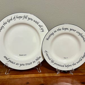 May include: Two white ceramic plates with black rims and inspirational text. The larger plate reads "May the God of hope fill you with all joy and peace as you trust in Him." The smaller plate says "Resting in the hope, your confidence, of eternal life promised before the world began."