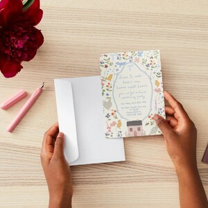May include: A floral invitation card with a house illustration is held near a white envelope. A pink pen and a red flower are on a light wood surface. The invitation has text within a floral border.