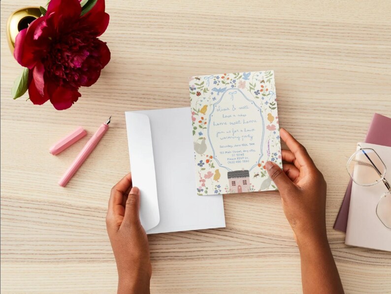 May include: A floral invitation card with a house illustration is held near a white envelope. A pink pen and a red flower are on a light wood surface. The invitation has text within a floral border.
