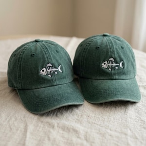 May include: Two dark green baseball caps with a washed-out finish. Each cap features an embroidered white and black fish skeleton graphic centred on the front panel. The hats have a curved brim, eyelets for ventilation, and a structured crown.