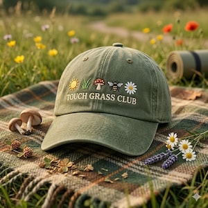 May include: Olive green baseball cap with embroidered sun, grass, mushroom, bee, and daisy graphics. The text "TOUCH GRASS CLUB" is embroidered below the graphics. The cap is resting on a plaid blanket with mushrooms and flowers.