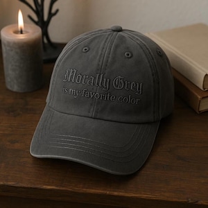 May include: A charcoal grey baseball cap with the embroidered text "Morally Grey is my favorite color." The cap has a curved brim and a button on top. The hat is sitting on a wooden surface.