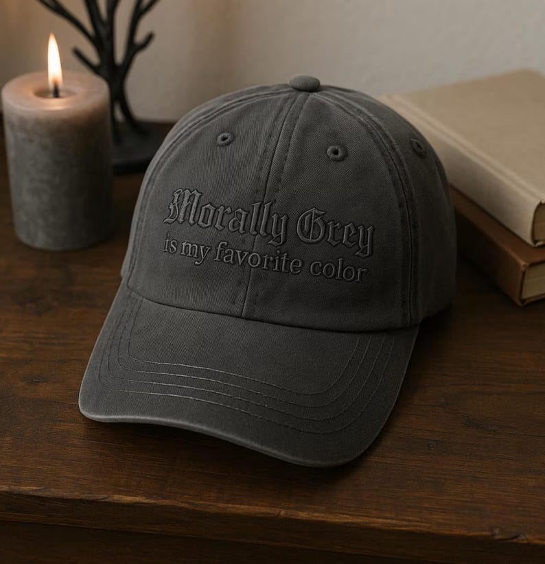 May include: A charcoal grey baseball cap with the embroidered text "Morally Grey is my favorite color." The cap has a curved brim and a button on top. The hat is sitting on a wooden surface.