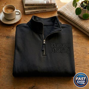 May include: Black quarter-zip sweatshirt with the embroidered phrase "Probably Nothing". The sweatshirt features a silver zip and is made of a soft material. The image also shows a coffee cup, books, and a small plant.