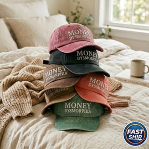 May include: A stack of six baseball caps in various colors, including dusty rose, black, brown, orange, and green. Each cap features the text "MONEY DYSMORPHIA" in white lettering. The caps are stacked on a bed with a neutral-toned blanket.