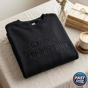 May include: Black crewneck sweatshirt with the embroidered text "Money Dysmorphia" in a gothic font. The sweatshirt is folded on a neutral-colored surface. A coffee cup and books are in the background.