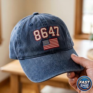Könnte beinhalten: Eine blaue Baseballkappe aus Denim mit der Zahl "8647" in Weiß mit roter Umrandung und einem kleinen amerikanischen Flaggenaufnäher. Die Mütze wird von einer Person gehalten.