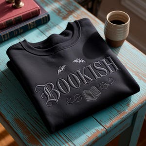 May include: Black crewneck sweatshirt with the word "BOOKISH" embroidered in gray script, with bat and book graphics. The sweatshirt is folded on a distressed teal table, with a coffee mug and books in the background.