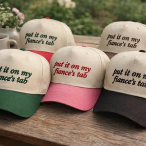 Puede incluir: Varias gorras de béisbol con la frase "put it on my fiance's tab" bordada. Las gorras tienen coronas de color crema y viseras de colores en verde, rojo, rosa y negro. El texto coincide con el color de la visera.