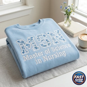 May include: A light blue sweatshirt with the letters "MSN" embroidered in white with a floral pattern. The words "Master of Science in Nursing" are embroidered below. A coffee cup and flowers are in the background.
