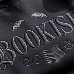 May include: Dark gray sweatshirt with the word "BOOKISH" embroidered in a gothic font. The design includes bat silhouettes and an open book illustration, creating a literary and spooky theme.