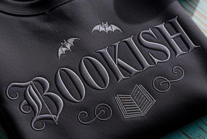 May include: Dark gray sweatshirt with the word "BOOKISH" embroidered in a gothic font. The design includes bat silhouettes and an open book illustration, creating a literary and spooky theme.