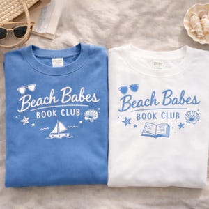 Può includere: Due felpe girocollo, una blu e una bianca, con la scritta "Beach Babes Book Club". Il design include occhiali da sole, una conchiglia, una barca a vela e stelle. La felpa blu è a tinta unita, mentre quella bianca ha un design simile.