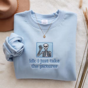 May include: Pale blue sweatshirt with a crew neck and long sleeves. The front has an embroidered patch with a skeleton waving and the text "idk i just take the pictures". A gold necklace with a small pendant is also visible.