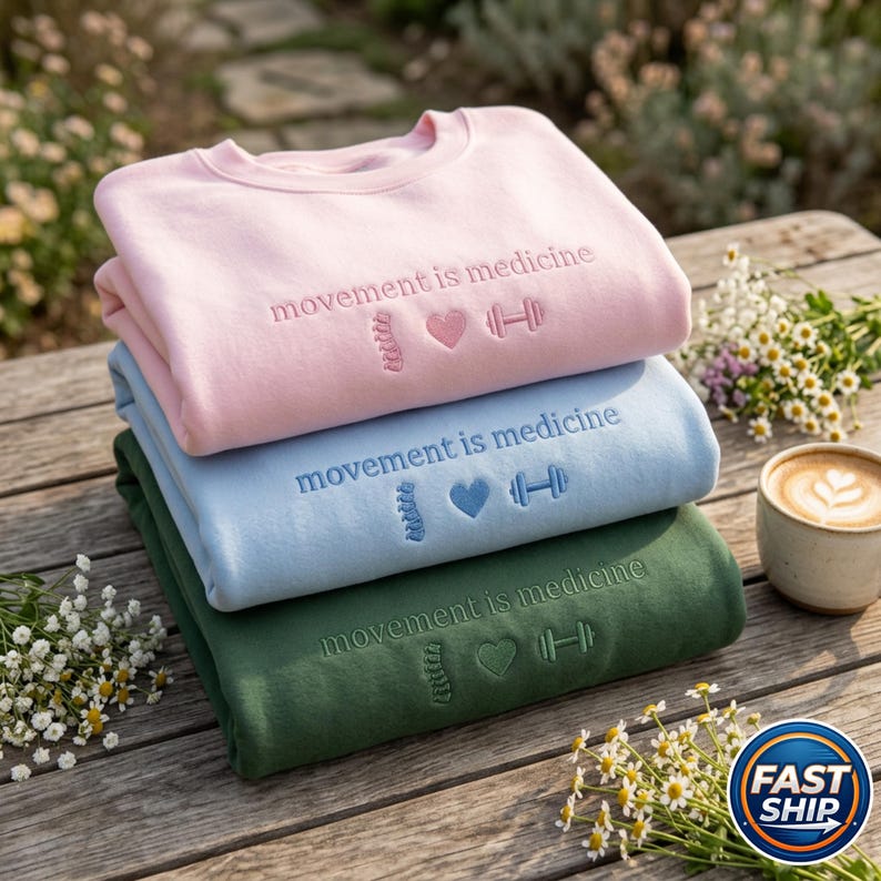 Peut inclure: Trois sweatshirts empil&eacute;s, rose, bleu et vert, brod&eacute;s avec "movement is medicine" et des symboles. Ils sont pos&eacute;s sur une table en bois, avec des fleurs et une tasse de caf&eacute;.