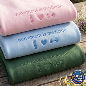 Peut inclure: Trois sweat-shirts empil&eacute;s, rose, bleu et vert. Chaque sweat-shirt porte la broderie "movement is medicine" avec un dessin de colonne vert&eacute;brale, c&oelig;ur et halt&egrave;re. Les sweat-shirts sont pli&eacute;s proprement.