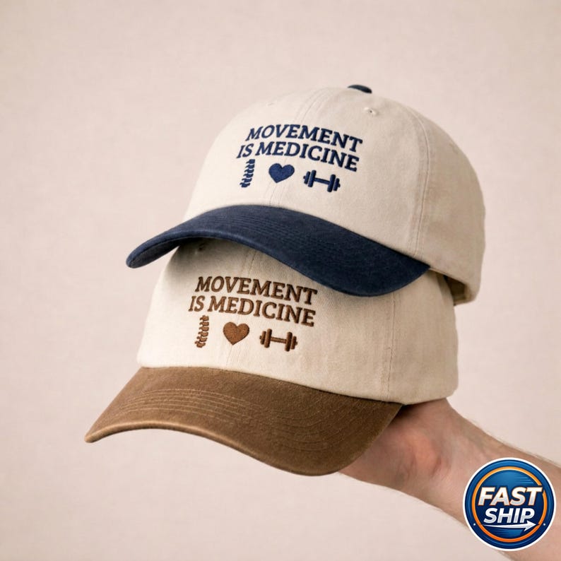 May include: Two baseball caps stacked on top of each other. The top cap has a navy blue brim, and the bottom cap has a brown brim. Both caps are beige with the embroidered text "MOVEMENT IS MEDICINE" and a heart, spine, and dumbbell graphic.