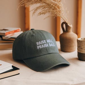 Puede incluir: Gorra de béisbol verde oscuro con el texto bordado "RAISE HELL, PRAISE DALE" en blanco. La gorra está sobre una superficie de color claro, con un jarrón y libros en el fondo.
