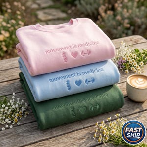 Peut inclure: Trois sweat-shirts empil&eacute;s, rose, bleu clair et vert fonc&eacute;, brod&eacute;s avec "movement is medicine", une colonne vert&eacute;brale, un c&oelig;ur et un halt&egrave;re. Ils sont pos&eacute;s sur une table en bois, avec des fleurs et une tasse de caf&eacute; &agrave; proximit&eacute;.