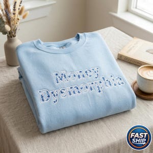 May include: A light blue sweatshirt with the embroidered text "Money Dysmorphia" in white with blue floral accents. The folded sweatshirt is on a neutral-toned surface, with a vase of dried flowers and a cup of coffee in the background.