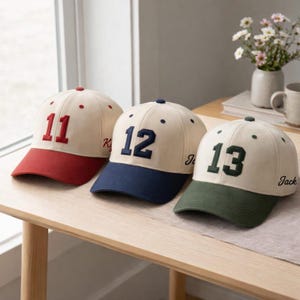 Puede incluir: Tres gorras de béisbol con coronas color crema y viseras de colores. Cada gorra tiene un número diferente bordado en la parte delantera: 11 en rojo, 12 en azul marino y 13 en verde. Cada gorra también tiene un nombre bordado en el lateral.