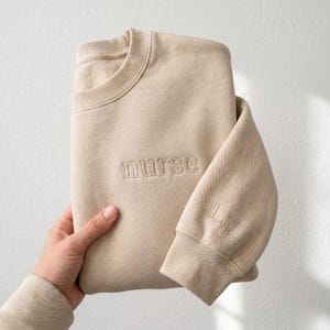 Puede incluir: Sudadera beige con la palabra "nurse" bordada en el pecho. La manga presenta iniciales y un estetoscopio. La prenda está doblada, mostrando su diseño y color.