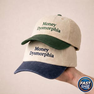 May include: Two baseball caps stacked on top of each other. The top cap has a tan crown and a dark green brim. The bottom cap has a tan crown and a navy blue brim. Both caps have the words "Money Dysmorphia" embroidered on the front.