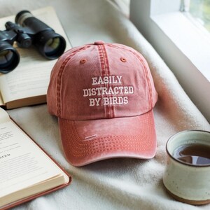 May include: A distressed red baseball cap with the text "EASILY DISTRACTED BY BIRDS" embroidered in white. The cap is sitting next to a cup of tea, binoculars, and an open book.