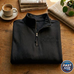 May include: Black quarter-zip sweatshirt with the embroidered text "Money Dysmorphia." The sweatshirt has a silver zipper and a fleece lining. The image is on a wooden surface, with a cup of coffee and books in the background.