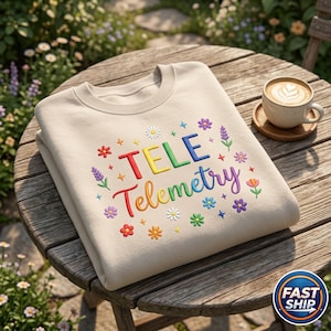 Peut inclure: Sweat-shirt beige avec l'inscription "TELE Telemetry" brodée en couleurs arc-en-ciel, entourée de motifs floraux colorés. Le sweat-shirt plié est posé sur une table en bois, avec une tasse de café en arrière-plan.