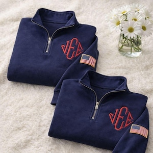 Könnte beinhalten: Zwei marineblaue Sweatshirts mit Viertelreißverschluss, roten gestickten Initialen und einem amerikanischen Flaggenaufnäher auf dem Ärmel. Die Sweatshirts sind auf einer weißen, strukturierten Oberfläche gefaltet, mit einer Vase mit weißen Gänseblümchen im Hintergrund.