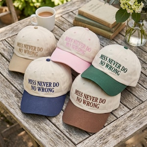 Op de afbeelding: Een verzameling baseballpetten in beige, roze, marineblauw, groen en bruin. Elke pet is geborduurd met de tekst "MISS NEVER DO NO WRONG". De petten liggen op een houten tafel en bieden een verscheidenheid aan kleuropties.