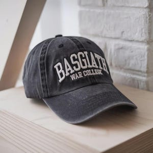 May include: A dark gray baseball cap with the text "BASGIATH WAR COLLEGE" embroidered in white. The cap has a curved brim and a distressed, vintage look, sitting on a wooden surface.