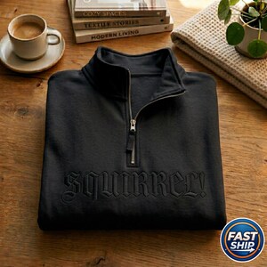 May include: Black sweatshirt with a quarter-zip collar and the word "SQUIRREL!" embroidered across the chest. The sweatshirt is folded on a wooden surface, with a cup of coffee and books in the background.