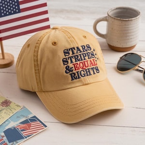Puede incluir: Gorra de béisbol color mostaza con el texto bordado "STARS STRIPES & EQUAL RIGHTS" en azul marino y rojo. La gorra está sobre una superficie de madera blanca, con una pequeña bandera americana y una taza en el fondo.