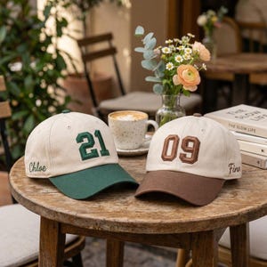 Puede incluir: Dos gorras de béisbol personalizadas sobre una mesa de madera. Una gorra es beige con visera verde y el número "21" bordado en verde. La otra gorra es beige con visera marrón y el número "09" bordado en marrón.