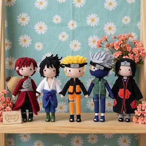 May include: Five handmade crochet dolls representing anime characters. The figures feature distinct outfits including a red coat, blue and white attire, an orange jumpsuit, a green vest with blue pants, and a black cloak with red cloud patterns.