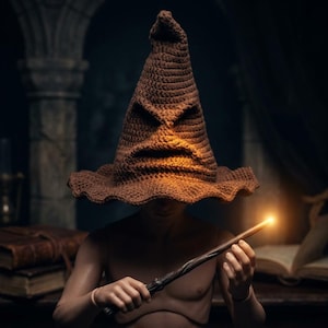 May include: A brown crocheted pointed hat with a wide, wavy brim and a textured, face-like appearance featuring deep folds for eyes and a mouth. A thin, dark brown wand with a glowing, bright light at the tip is held in front of the chest.
